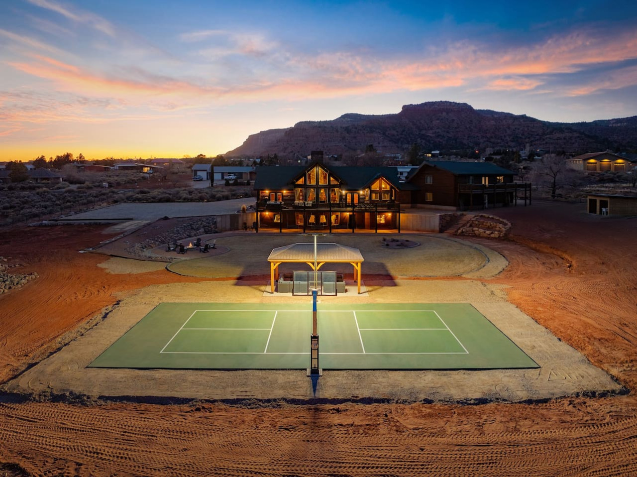 The Blackhawk Cabin in Kanab at dusk, with a lit pickleball court and outdoor movie pavilion in front of the red cliffs